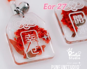 May include: A clear resin charm with a red dried flower and white Chinese characters that read "Happiness" in a white outline. The charm is attached to a silver bell and a red beaded chain.