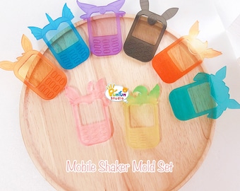 May include: A set of eight clear plastic phone-shaped molds for making shaker charms. The molds are in various colors, including purple, gray, orange, blue, pink, yellow, and teal. The molds are arranged on a wooden surface. The text "Mobile Shaker Mold Set" is printed on the surface.