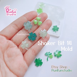 Puede incluir: Un molde de silicona transparente con cuatro formas de trébol. El molde está etiquetado como "Shaker Bit 14 Mold". También hay varios tréboles de resina terminados en diferentes colores, incluyendo verde, verde claro y blanco.