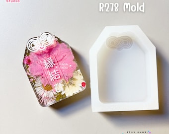 Puede incluir: Un molde de silicona blanco para fundición de resina, con forma de etiqueta con la parte superior redondeada y un pequeño símbolo de infinito en la parte superior. El molde se muestra junto a una pieza de resina terminada con flores prensadas y el texto "福" en blanco.