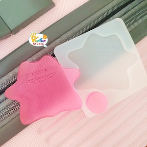 May include: Two pink star-shaped silicone luggage tags with a cartoon face. The tag on the left is pink and the tag on the right is translucent.