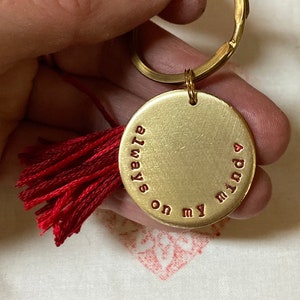 May include: A gold-colored keychain with a round pendant. The pendant has the words "always on my mind" in red lettering. A red tassel hangs from the pendant. The keychain is held in a hand.