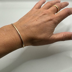 Può includere: Un braccialetto sottile color oro e un anello abbinato. Il braccialetto è una semplice fascia continua. L'anello è una fascia sottile. Entrambi i pezzi sono esposti su una mano su sfondo bianco. Gioielli.