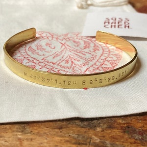 Puede incluir: Una pulsera de puño dorada con coordenadas grabadas. La pulsera está sobre una tela blanca con un patrón rojo. Una pequeña etiqueta con el texto "swiss made chen" es visible en el fondo.