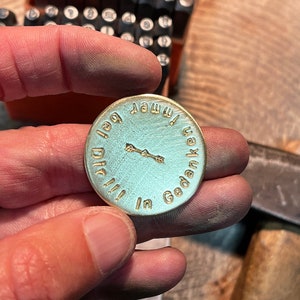 May include: A round, light blue metal pendant with gold-colored text and a small arrow design. The text reads "in Gedanken immer bei Dir!!!". The pendant is held in a hand, with metal stamping tools in the background.