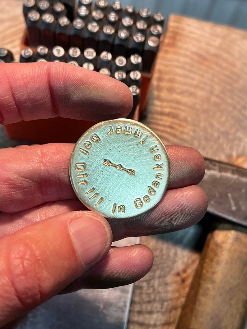 May include: A round, light blue metal pendant with gold-colored text and a small arrow design. The text reads "in Gedanken immer bei Dir!!!". The pendant is held in a hand, with metal stamping tools in the background.