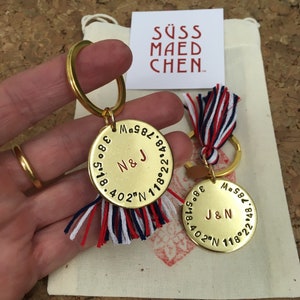 Könnte beinhalten: Zwei goldene Schlüsselanhänger mit rot-weiß-blauen Quasten. Jeder Schlüsselanhänger hat ein rundes Metallschild mit den Initialen "N & J" und "J & N", die in Rot graviert sind. Die Schilder haben auch Zahlen und Koordinaten, die um den Rand graviert sind.