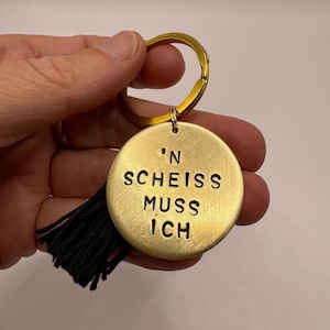 Könnte beinhalten: Ein runder Schlüsselanhänger aus gebürstetem goldfarbenem Metall mit dem eingestanzten schwarzen Text 'N SCHEISS MUSS ICH. Der kreisförmige Anhänger ist an einem goldfarbenen Schlüsselring befestigt und hat eine kleine schwarze Stoffquaste.