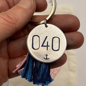 May include: A silver keyring with a circular pendant displaying the number "040" and an anchor symbol. It has a blue and pink tassel. The keyring is held by a hand.