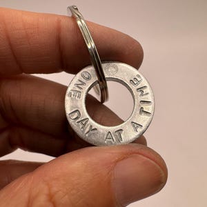 Pode incluir: Um porta-chaves circular em metal prateado com o texto estampado ONE DAY AT A TIME e um pequeno ícone de sol. O pendente está preso a uma argola de metal prateado padrão, criando um acessório funcional para chaves ou malas.