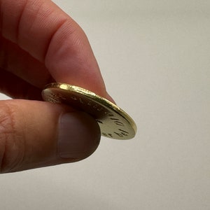 May include: A gold-colored coin held between fingers. The coin's edge is textured, and the surface has engraved text. The coin appears to be slightly worn, suggesting it may be an older piece.