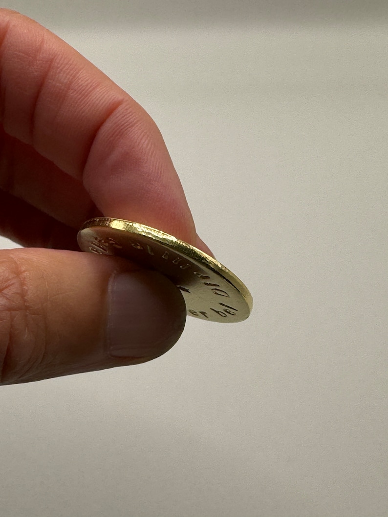 May include: A gold-colored coin held between fingers. The coin's edge is textured, and the surface has engraved text. The coin appears to be slightly worn, suggesting it may be an older piece.