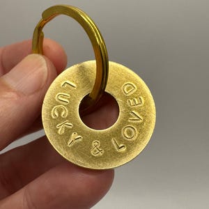 Könnte beinhalten: Ein runder Schlüsselanhänger aus goldfarbenem Metall mit gebürsteter Oberfläche und einem Loch in der Mitte. Der Text LUCKY & LOVED ist in die Oberfläche der Metallscheibe eingeprägt. Ein passender goldfarbener Metallring ist durch das Loch gezogen.