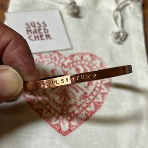 Könnte beinhalten: Ein kupferfarbenes Armband mit dem geprägten Schriftzug "SOUL SISTER". Das Armband wird vor einem weißen Kordelzugbeutel und einer Karte mit dem Text "SQSS MAED CHEN" gehalten. Im Hintergrund ist ein rotes Herzdesign zu sehen.