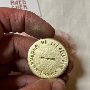 May include: A round, brass-colored pendant with the words "Immer bei Dir" and "Gedanken" encircling a central arrow design. The pendant is held in a hand, with a tag in the background that reads "SOSS MAEDCHEN".