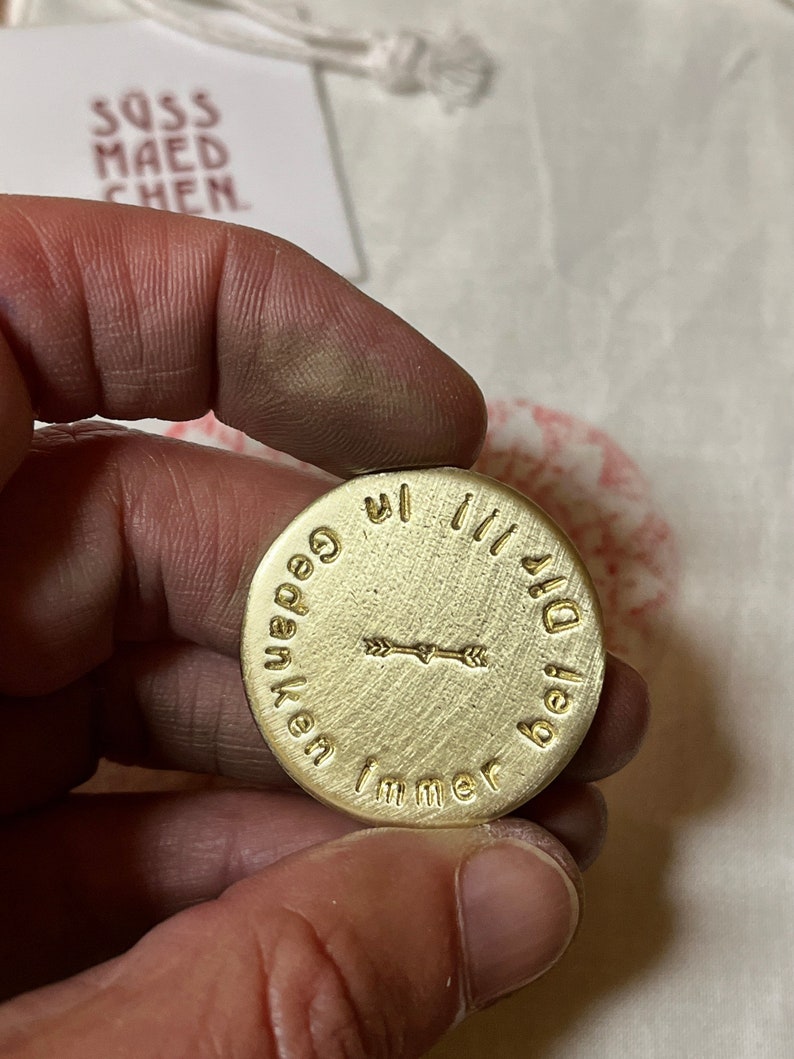 May include: A round, brass-colored pendant with the words "Immer bei Dir" and "Gedanken" encircling a central arrow design. The pendant is held in a hand, with a tag in the background that reads "SOSS MAEDCHEN".