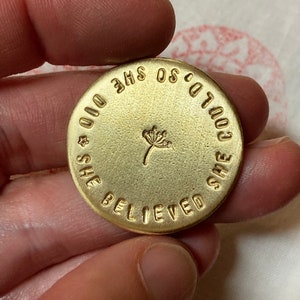 May include: A round, gold-colored metal coin with the stamped text "SHE BELIEVED SHE COULD SO SHE DID." A small dandelion design is in the center. The coin is held between fingers.