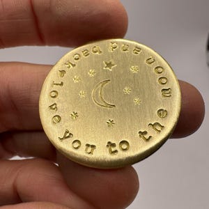 Könnte beinhalten: Ein kreisförmiger, goldfarbener Metall-Token mit einem eingravierten Halbmond in der Mitte, umgeben von kleinen Sternmotiven. Der äußere Rand ist mit dem Text "love you to the moon and back" in einem kreisförmigen Muster und einem Herz versehen.