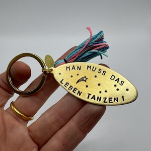 Könnte beinhalten: Ein goldfarbener Metall-Schlüsselanhänger mit dem deutschen Satz "MAN MUSS DAS LEBEN TANZEN!" auf einem ovalen Anhänger. Der Schlüsselanhänger hat einen bunten Quastenanhänger und einen goldenen Ring.