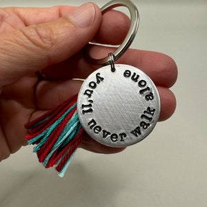 Op de afbeelding: Zilveren sleutelhanger met een rode en turquoise kwast. De sleutelhanger heeft een ronde metalen hanger met de tekst "You'll never walk alone" erop gegraveerd.