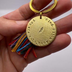 Puede incluir: Llavero dorado con una etiqueta redonda que dice "You are gold, baby.  P.I.T.O.S." La etiqueta tiene un símbolo de rayo y un borla de colores.