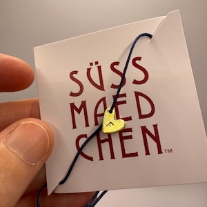 Könnte beinhalten: Eine weiße Karte mit dem Text "SÜSS MAEDCHEN" in roten Buchstaben und einem goldenen Herz-Anhänger, der an einer blauen Schnur befestigt ist. Die Karte hat ein kleines "TM"-Symbol in der unteren rechten Ecke.
