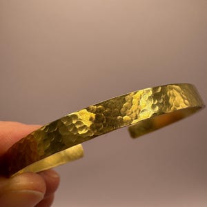 Könnte beinhalten: Ein goldfarbenes, gehämmertes Metall-Armband. Das Armband besteht aus dickem, strukturiertem Metall und hat ein schlichtes, elegantes Design.