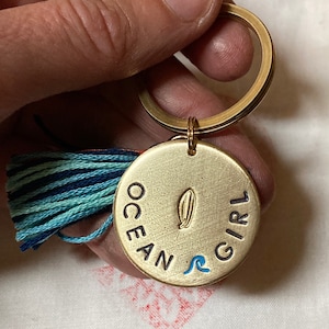 Könnte beinhalten: Ein goldfarbener Schlüsselanhänger mit einem runden Anhänger mit der Aufschrift "OCEAN GIRL". Der Anhänger zeigt ein Surfbrett und ein blaues Wellenmuster. Ein blau-türkisfarbener Quastenanhänger hängt am Schlüsselanhänger.