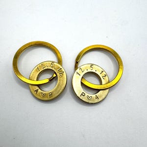 Könnte beinhalten: Zwei goldfarbene Schlüsselanhänger mit kreisförmigen Ringen und gravierten Unterlegscheiben. Jede Unterlegscheibe trägt das Datum "12.5.12", die Initialen "A P" und ein Herzsymbol. Die Schlüsselanhänger sind wahrscheinlich personalisierte Geschenke.