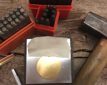 May include: A close-up of a metal stamping set with a variety of letter and number stamps. The stamps are arranged in a red plastic case. A small, round, brass disc is sitting on a black metal surface. A hammer is also visible in the image.