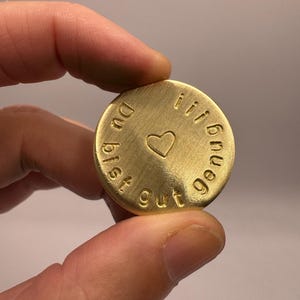Könnte beinhalten: Ein kreisförmiger, goldfarbener Metall-Token mit gebürsteter Oberfläche. Der Text "Du bist gut genug!!!" ist in einer serifenlosen Schrift am Rand eingeprägt, und ein kleines Herz-Symbol befindet sich mittig auf der Münze.