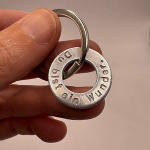 Könnte beinhalten: Ein silberfarbener Metallschlüsselanhänger mit einem runden, scheibenförmigen Anhänger. Der Anhänger ist mit dem deutschen Text 'Du bist ein wunder.' graviert. Der Schlüsselanhänger verfügt über einen Standard-Spaltring mit gebürsteter Oberfläche.