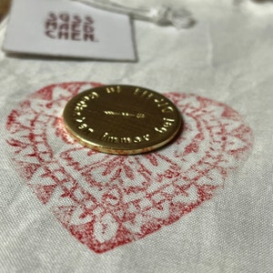 May include: A gold-colored coin with embossed text, resting on a white fabric pouch with a red heart-shaped design. A small tag with text is attached to the pouch. The coin's text is partially visible.