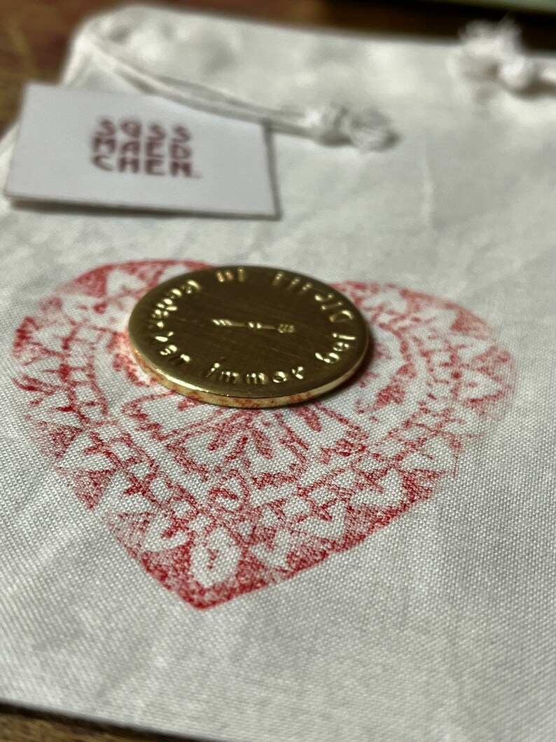 May include: A gold-colored coin with embossed text, resting on a white fabric pouch with a red heart-shaped design. A small tag with text is attached to the pouch. The coin's text is partially visible.