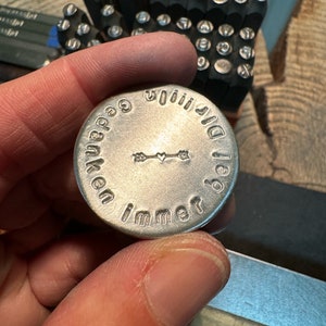May include: A silver metal disc with stamped text and an arrow design is held in a hand. The text around the edge reads "Gedanken immer bei dir". Letter stamps are visible in the background, along with a wooden surface.