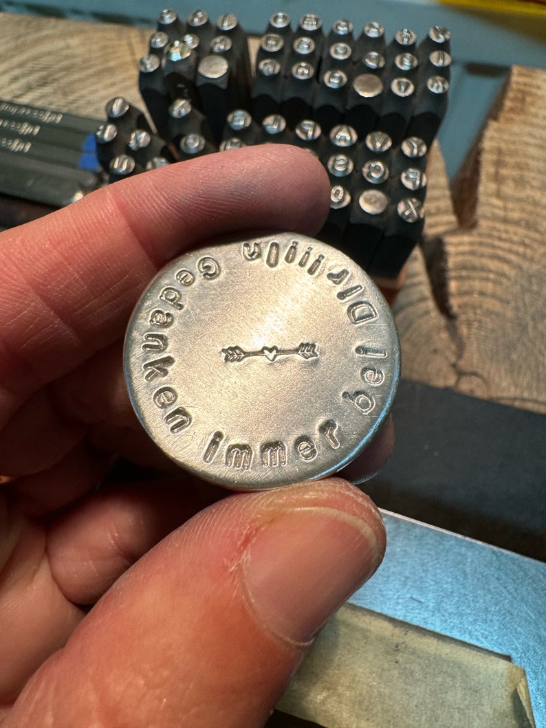 May include: A silver metal disc with stamped text and an arrow design is held in a hand. The text around the edge reads "Gedanken immer bei dir". Letter stamps are visible in the background, along with a wooden surface.