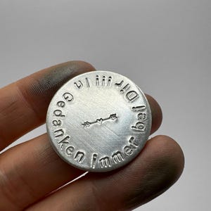 May include: A round, silver-colored metal pendant with embossed text and a small arrow design. The text encircles the edge of the pendant, with a heart-shaped arrow in the center. The pendant is held in a hand.