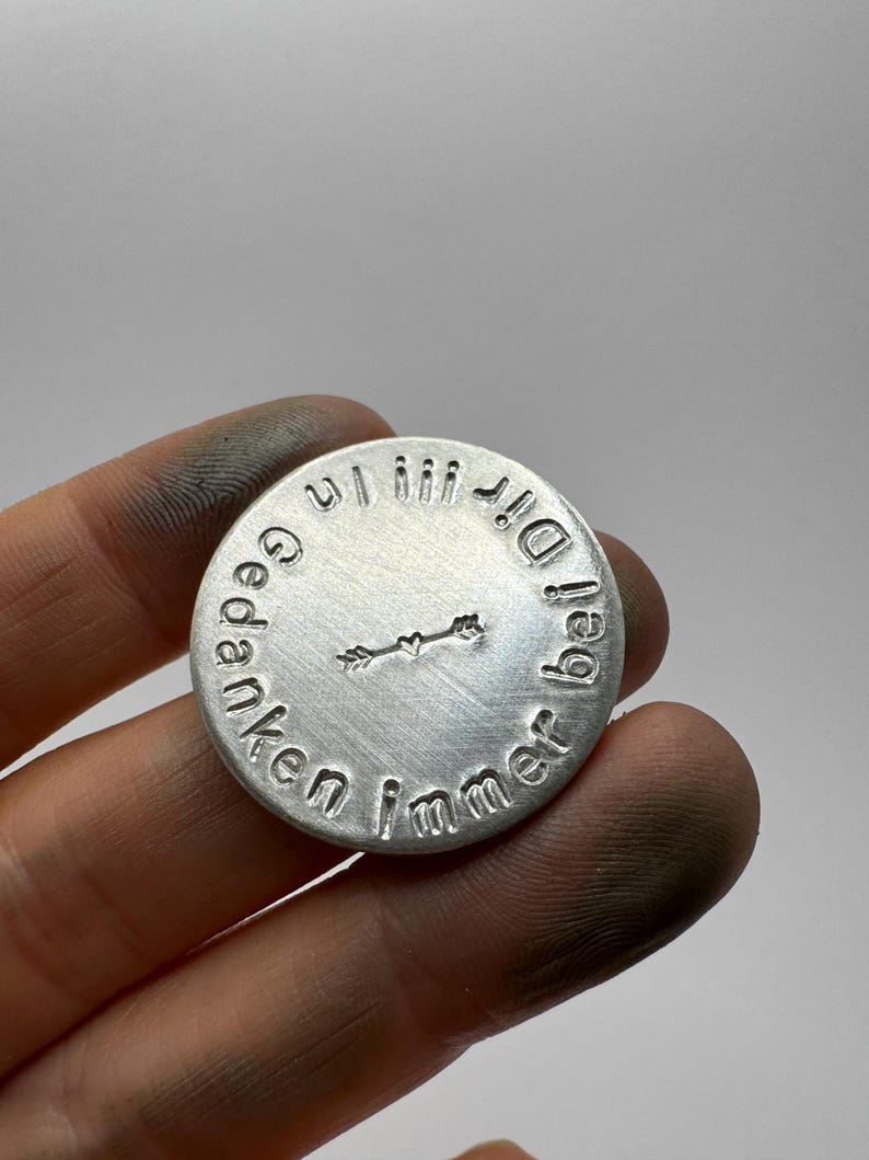 May include: A round, silver-colored metal pendant with embossed text and a small arrow design. The text encircles the edge of the pendant, with a heart-shaped arrow in the center. The pendant is held in a hand.