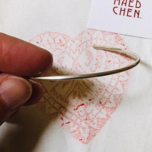 May include: A silver cuff bracelet held by a hand, set against a white background with a red heart-shaped design. A white card with the text "SÜSS MAEDCHEN" is also visible.