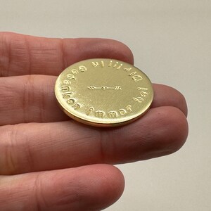 May include: A gold-colored, circular coin with embossed text and an arrow design. The coin is held in a hand, and the text appears to be in a foreign language. The coin's surface has a reflective sheen.