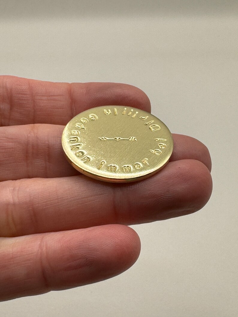 May include: A gold-colored, circular coin with embossed text and an arrow design. The coin is held in a hand, and the text appears to be in a foreign language. The coin's surface has a reflective sheen.