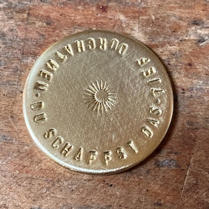 Könnte beinhalten: Eine goldfarbene Metallscheibe mit dem Text "DAS SCHAFFST DU - DURCHHALTEN" in einem Kreis um eine stilisierte Sonne.