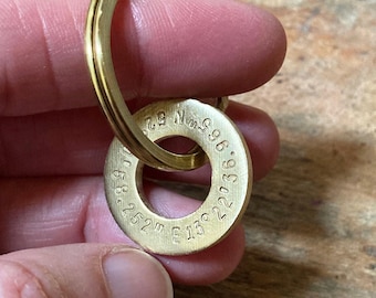 May include: A circular brass keychain charm featuring stamped geographic coordinates. The metal ring is polished and gold-toned, with the numbers 52 degrees 36.965 minutes North and 13 degrees 22.252 minutes East clearly engraved on the surface.