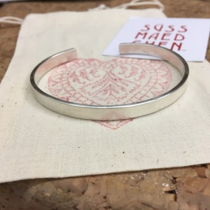 May include: A silver-colored cuff bracelet with a flat, rectangular design. The bracelet is resting on a cream-colored fabric pouch with a red heart-shaped design. A small white card with red text is also visible.