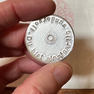 Puede incluir: Un sello de metal plateado con el texto "Du bist einzigartig.  Du  und  deine  voll."