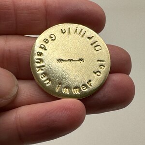 May include: A round, gold-colored metal coin with embossed text and arrow design. The text encircles the coin, with an arrow design in the center. The coin is held in a hand.