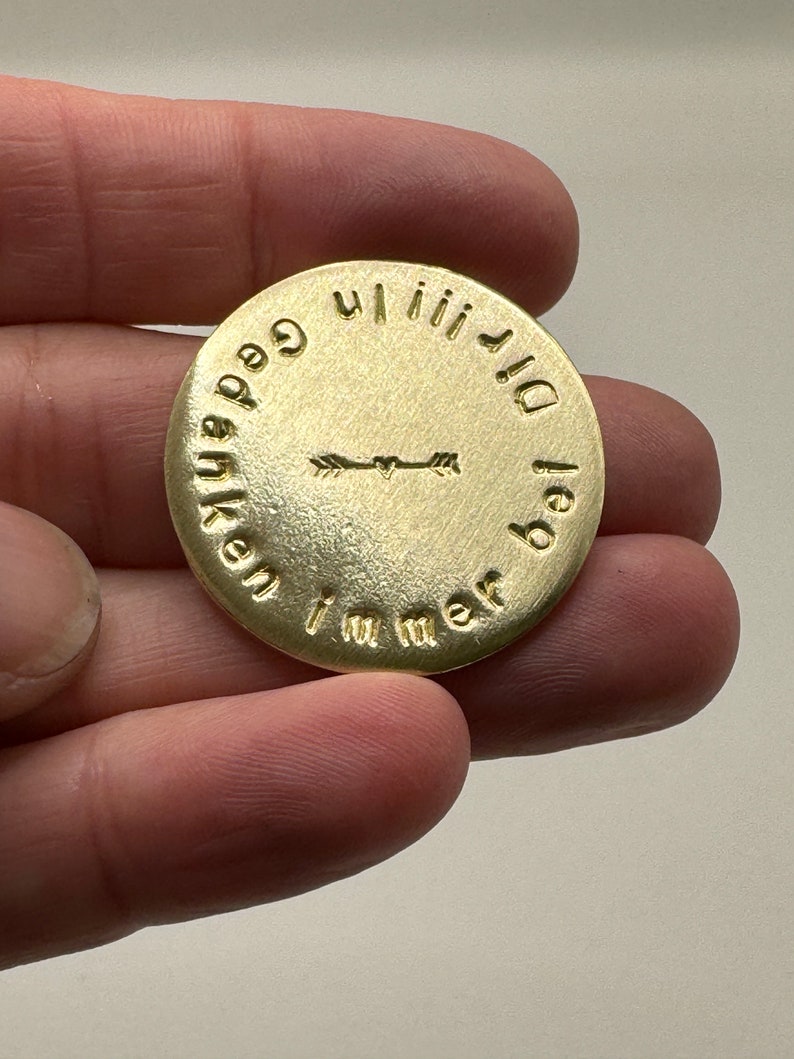 May include: A round, gold-colored metal coin with embossed text and arrow design. The text encircles the coin, with an arrow design in the center. The coin is held in a hand.
