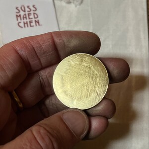May include: A round, brushed brass coin held in a hand. The coin has a textured surface with visible grain. A white tag with the text "S&Uuml;SS MAEDCHEN" is visible in the background.
