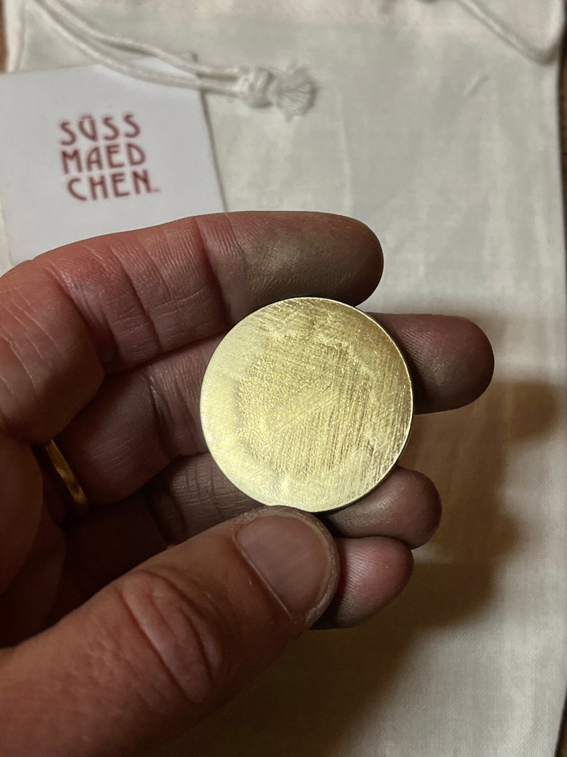 May include: A round, brushed brass coin held in a hand. The coin has a textured surface with visible grain. A white tag with the text "S&Uuml;SS MAEDCHEN" is visible in the background.