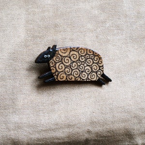 Könnte beinhalten: Eine Holzspange in Form eines Schafs mit schwarzem Gesicht und wirbelnden braunen Markierungen. Die Spange befindet sich auf einem beigefarbenen Canvas-Hintergrund.